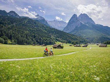 Hammersbach in Grainau, the Zugspitze in the background Two cyclists on a cycle path between fields and wooden huts, with the Zugspitze massif in the background